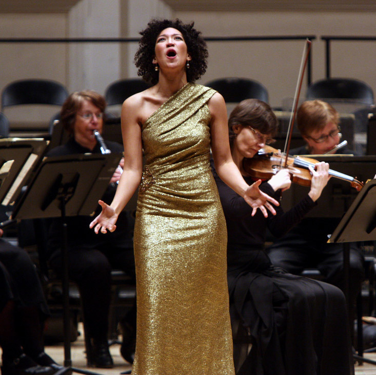 Julia Bullock wears a golden dress, singing in front of an orchestra with her arms outstretched.