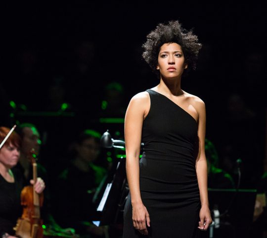 Julia stares seriously into the distance as she stands in front of an orchestra.