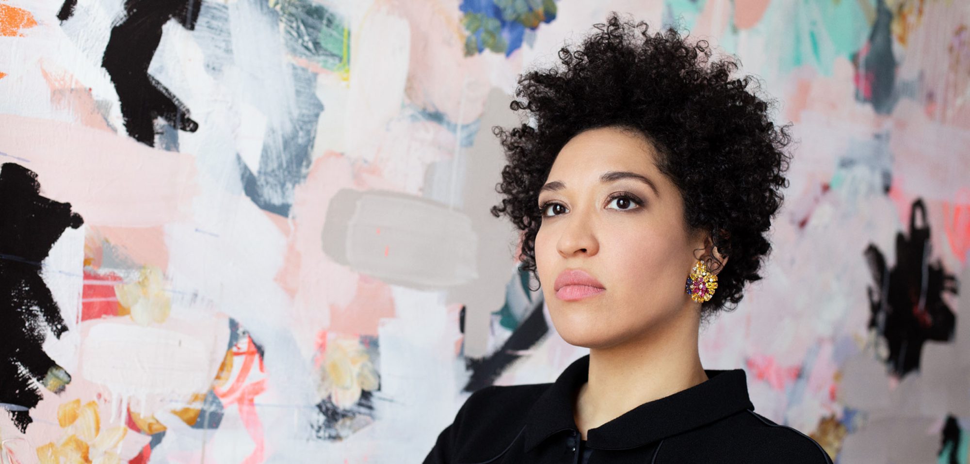 Julia Bullock wears a black blazer, looking of to the side seriously, standing against a backdrop of abstract shapes.