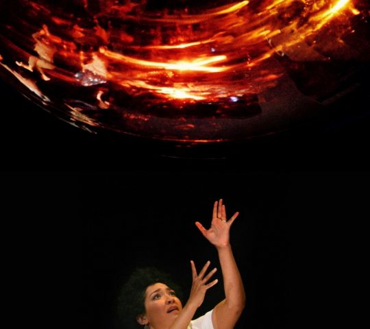 Julia Bullock performs on stage with her arms raised above her head, stretched towards a projection of what appears to be a light.
