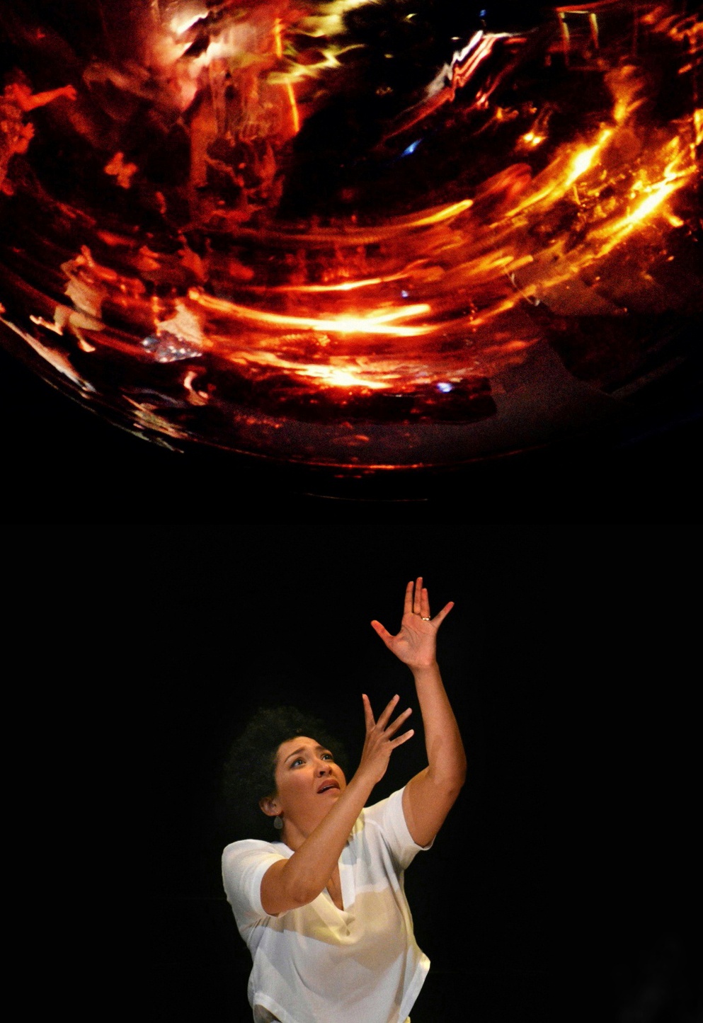 Julia Bullock performs on stage with her arms raised above her head, stretched towards a projection of what appears to be a light.