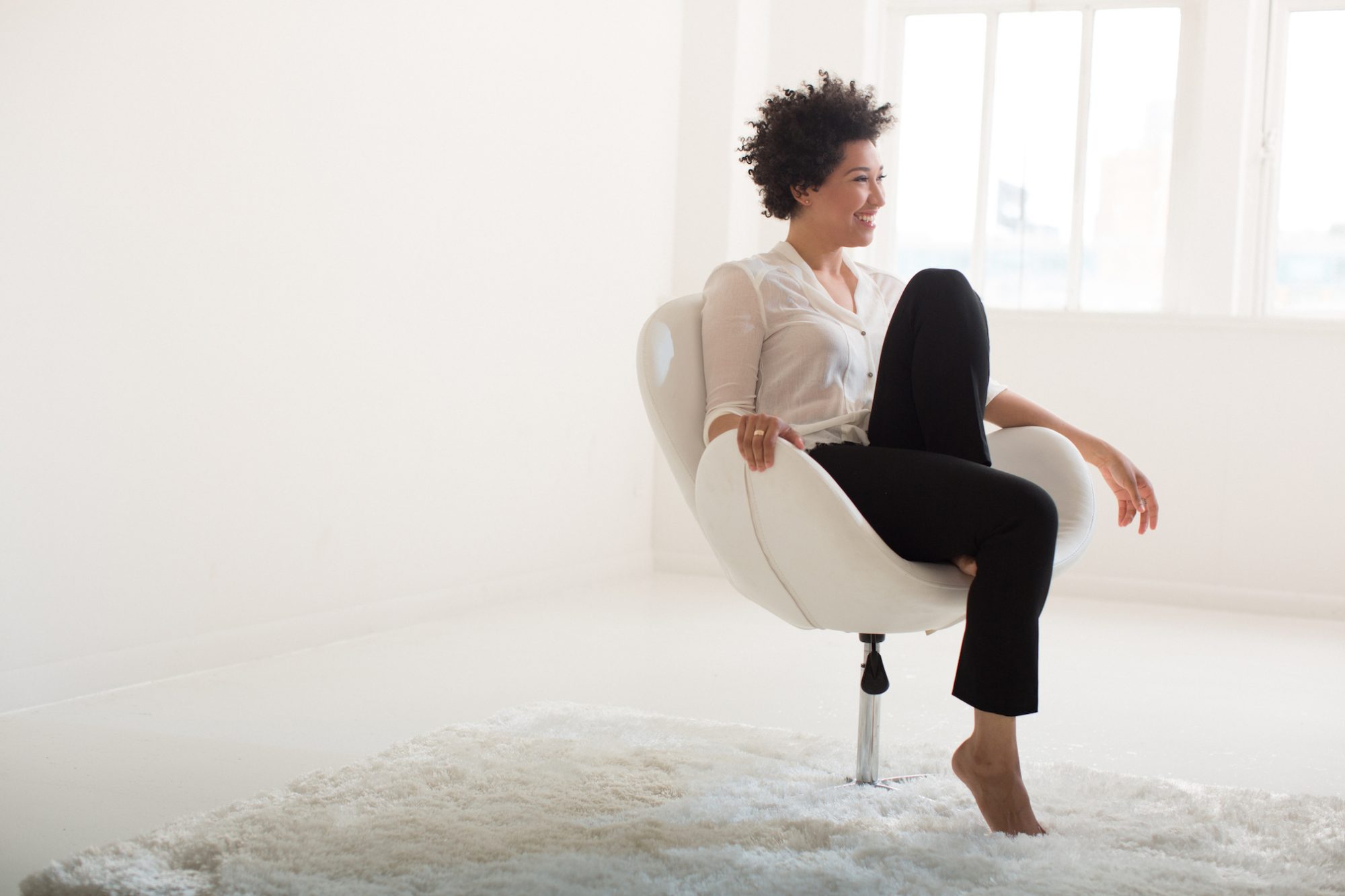 Julia Bullock sits in a white chair, wearing a white blouse and black pants, with one leg folded up on the chair. .