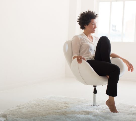 Julia Bullock sits in a white chair, wearing a white blouse and black pants, with one leg folded up on the chair. .