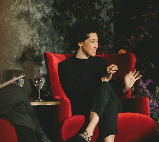 Julia Bullock sits on a stage in a red chair with one leg crossed over the other, smiling and making a descriptive motion with her hands.