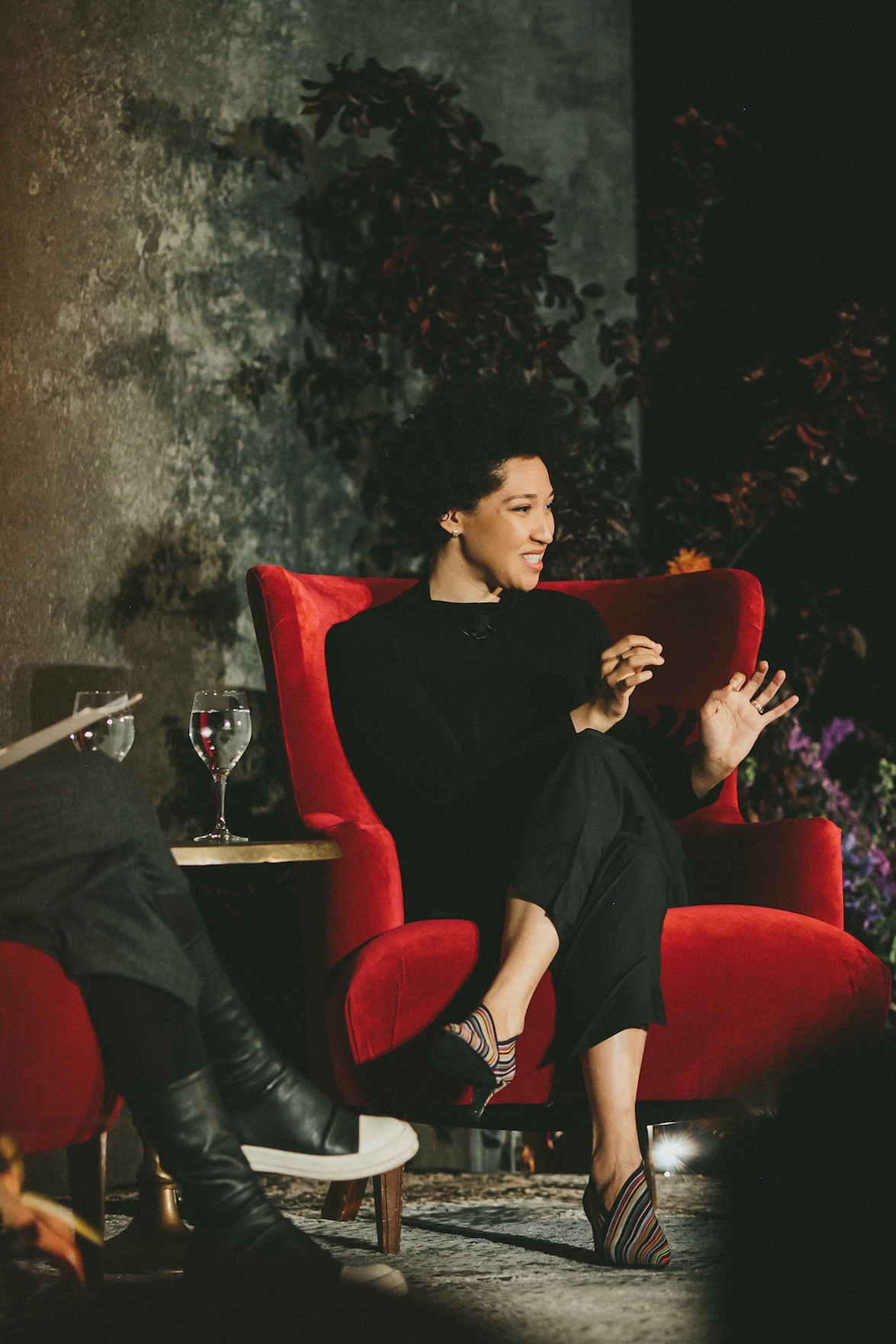 Julia Bullock sits on a stage in a red chair with one leg crossed over the other, smiling and making a descriptive motion with her hands.
