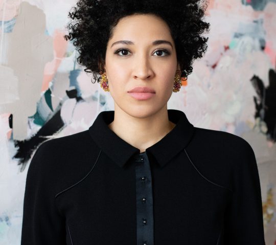 Julia Bullock wears a black blazer, looking straight forward seriously against a backdrop of abstract shapes.