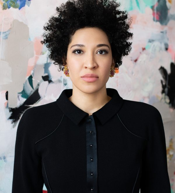 Julia Bullock wears a black blazer, looking straight forward seriously against a backdrop of abstract shapes.