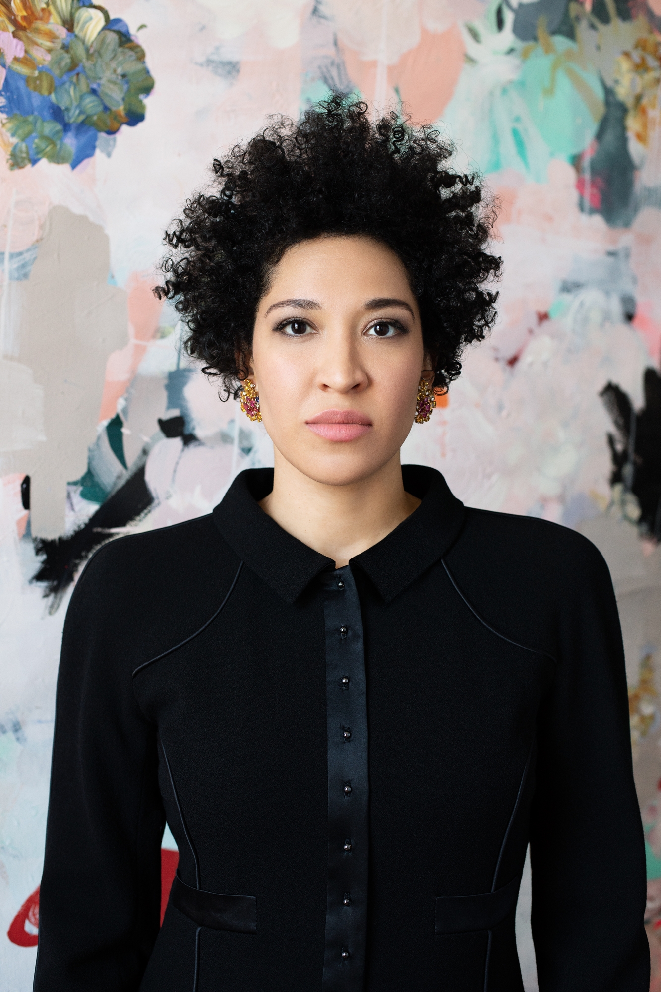 Julia Bullock wears a black blazer, looking straight forward seriously against a backdrop of abstract shapes.