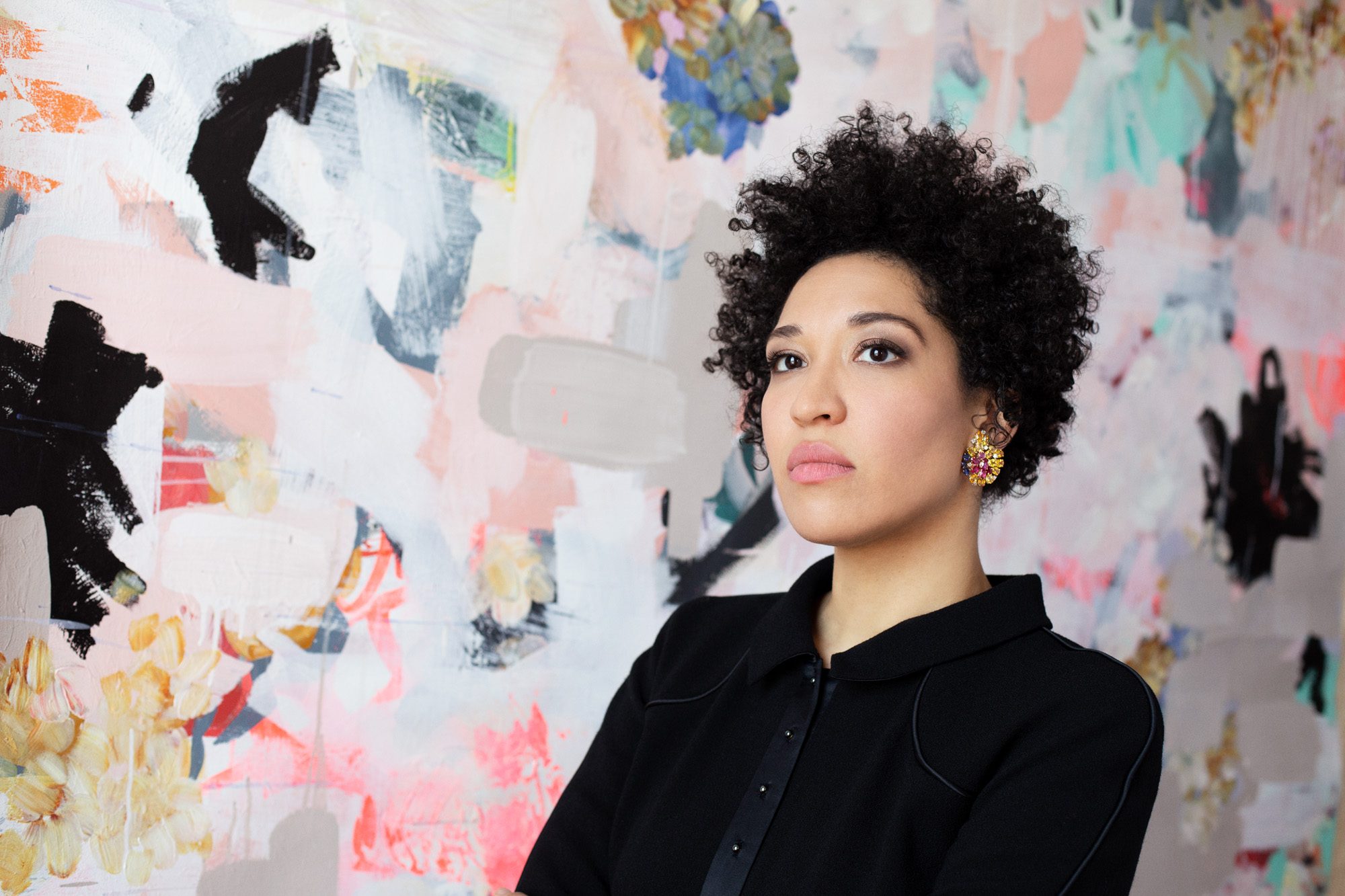 Julia Bullock wears a black blazer, looking of to the side seriously, standing against a backdrop of abstract shapes.