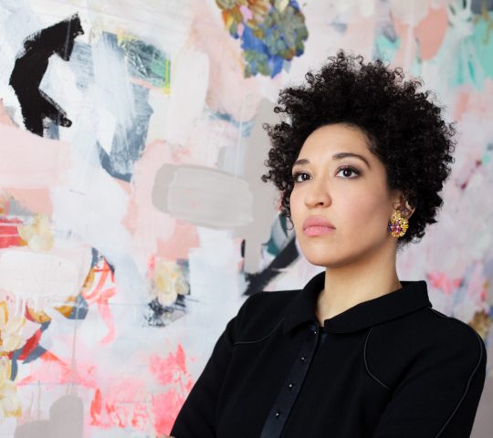 Julia Bullock wears a black blazer, looking of to the side seriously, standing against a backdrop of abstract shapes.