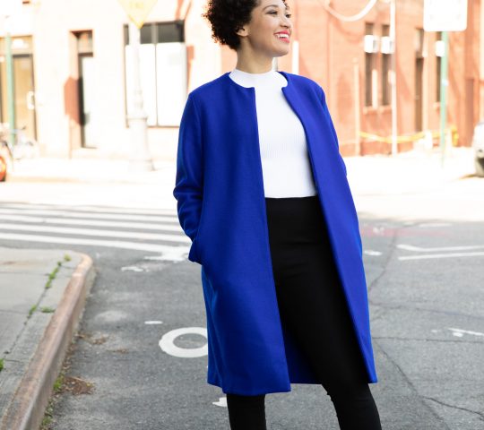 Julia Bullock stands on a street corner wearing a blue jacket, a white shirt, and black pants, smiling and looking to the side.