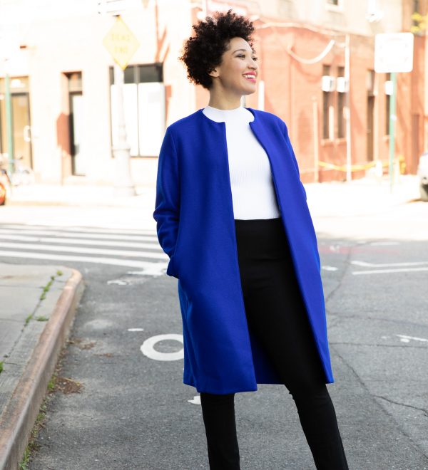Julia Bullock stands on a street corner wearing a blue jacket, a white shirt, and black pants, smiling and looking to the side.