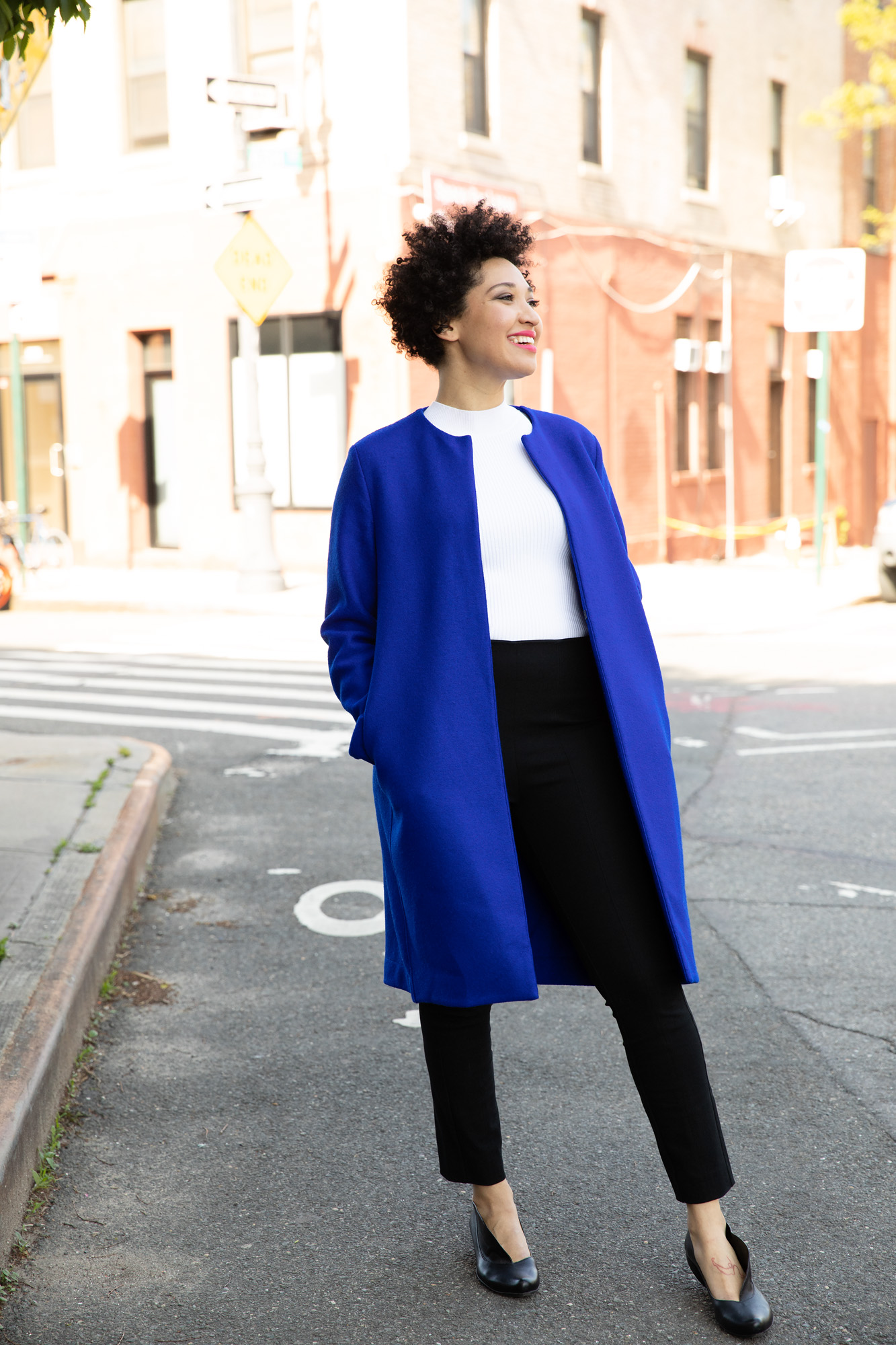 Julia Bullock stands on a street corner wearing a blue jacket, a white shirt, and black pants, smiling and looking to the side.