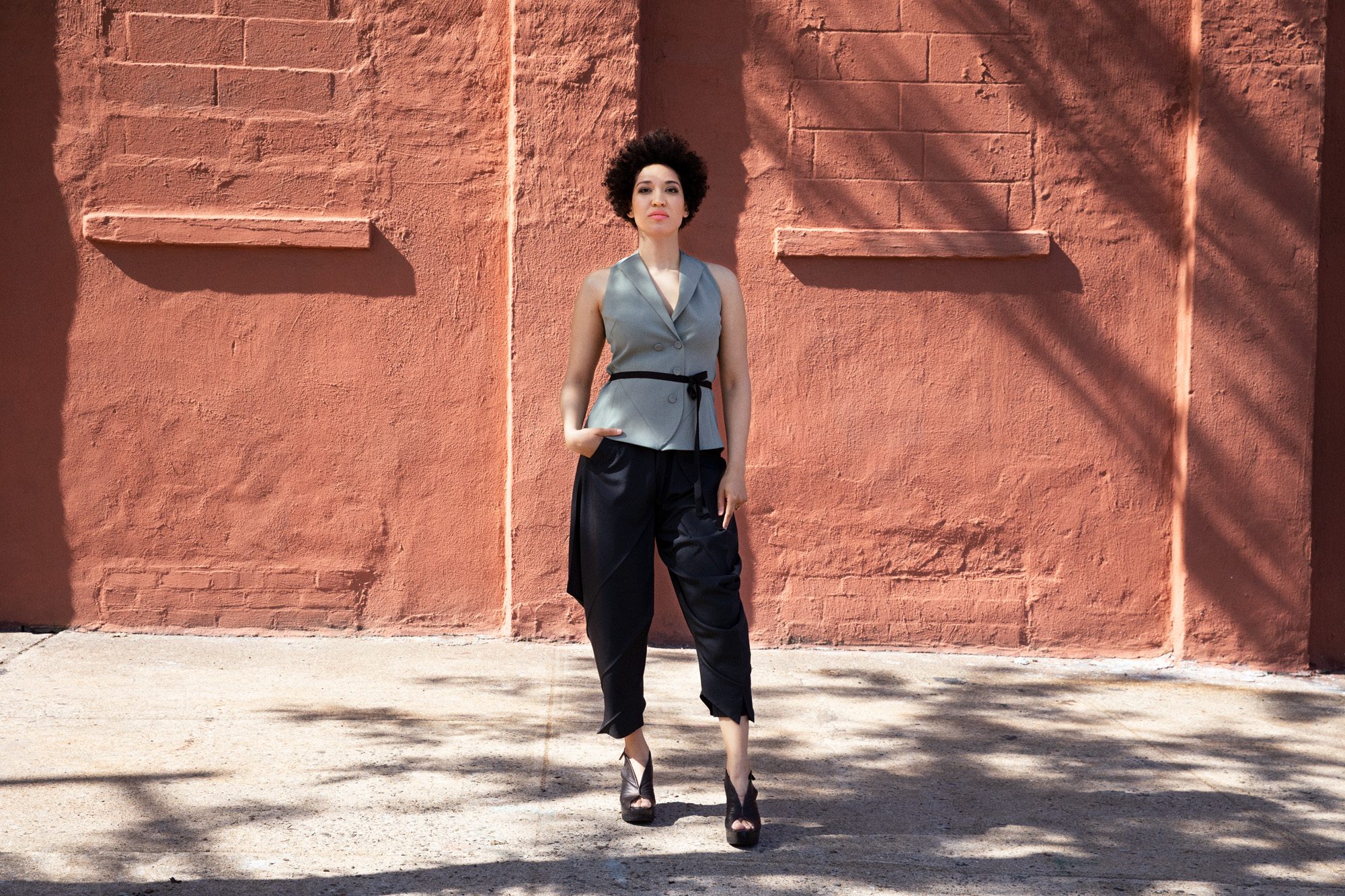 Julia Bullock stands in front of a red brick wall with one hand in her pocket wearing a grey vest with buttons and unstructured black pants.