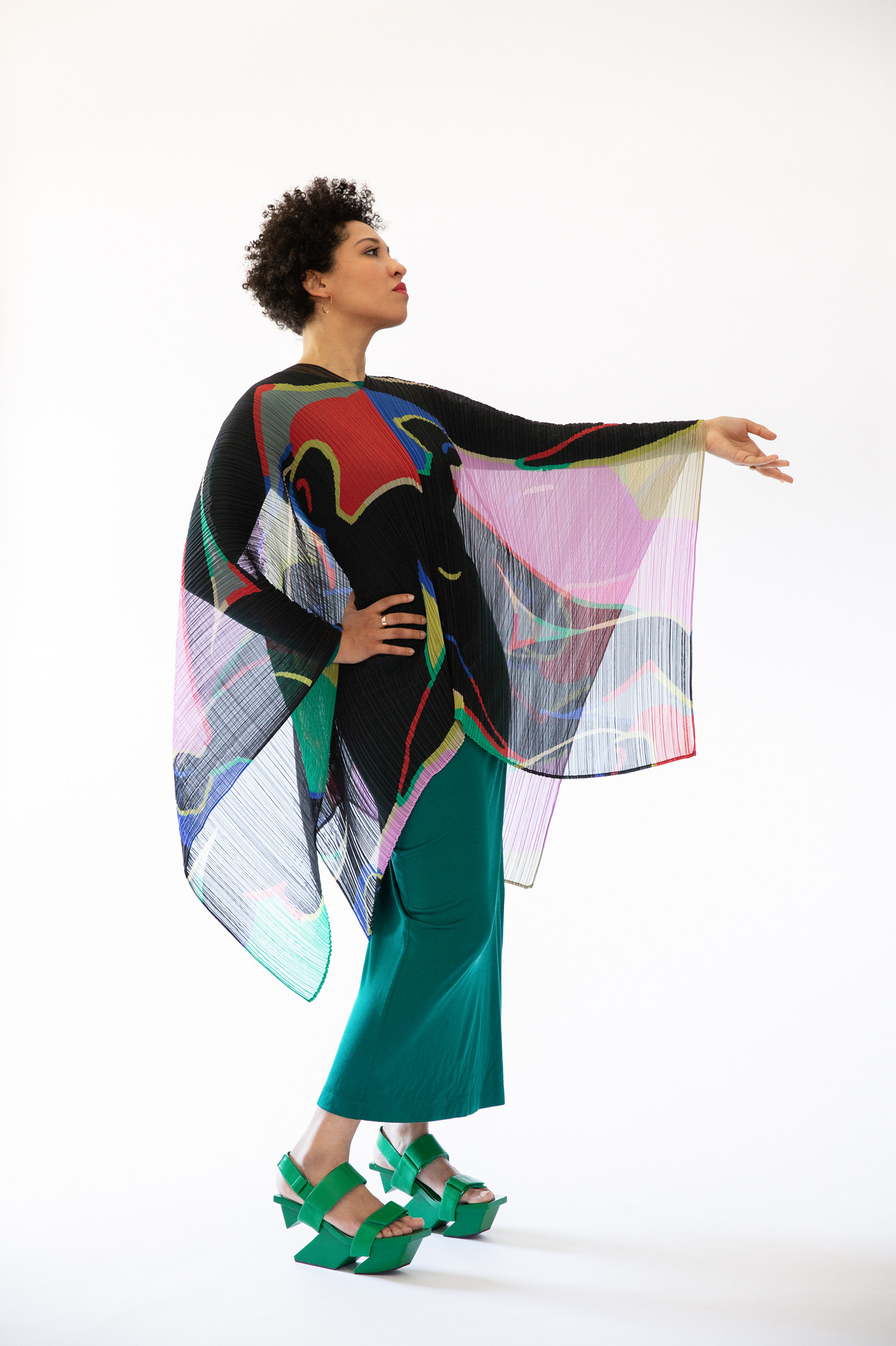 Julia Bullock wears an abstract patterned top with loose green pants. She rests one hand on her hip and extends the other arm out in front of her.