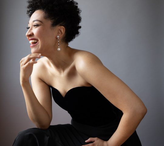 A profile of Julia Bullock, where she wears a black dress, pearl ring and earrings, leaning forward and smiling.
