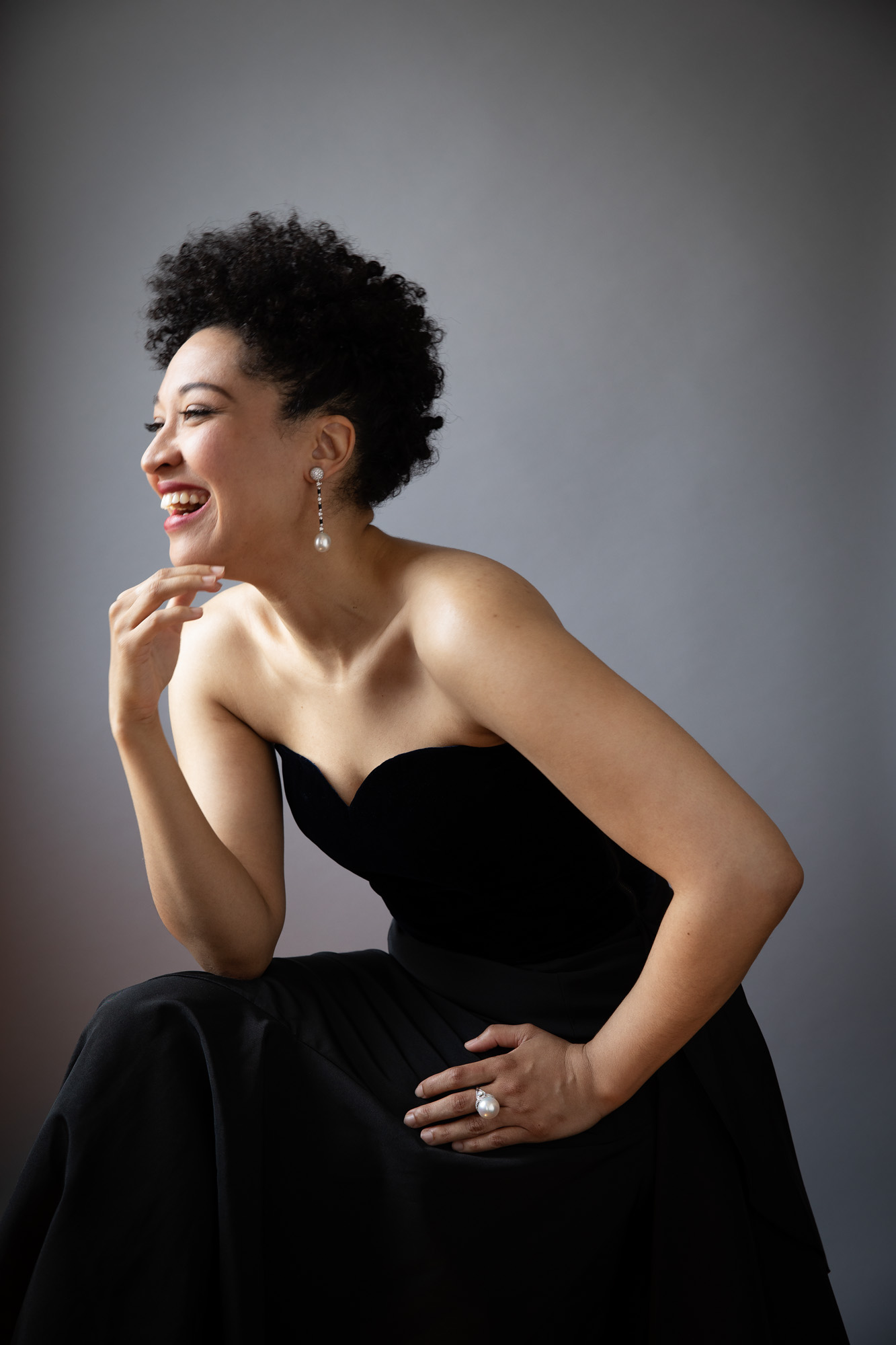 A profile of Julia Bullock, where she wears a black dress, pearl ring and earrings, leaning forward and smiling.