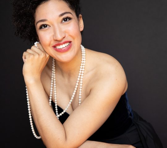 Julia Bullock wears a black sleeveless gown and a white pearl necklace as she leans forward, resting her cheek on her hand and smiling.