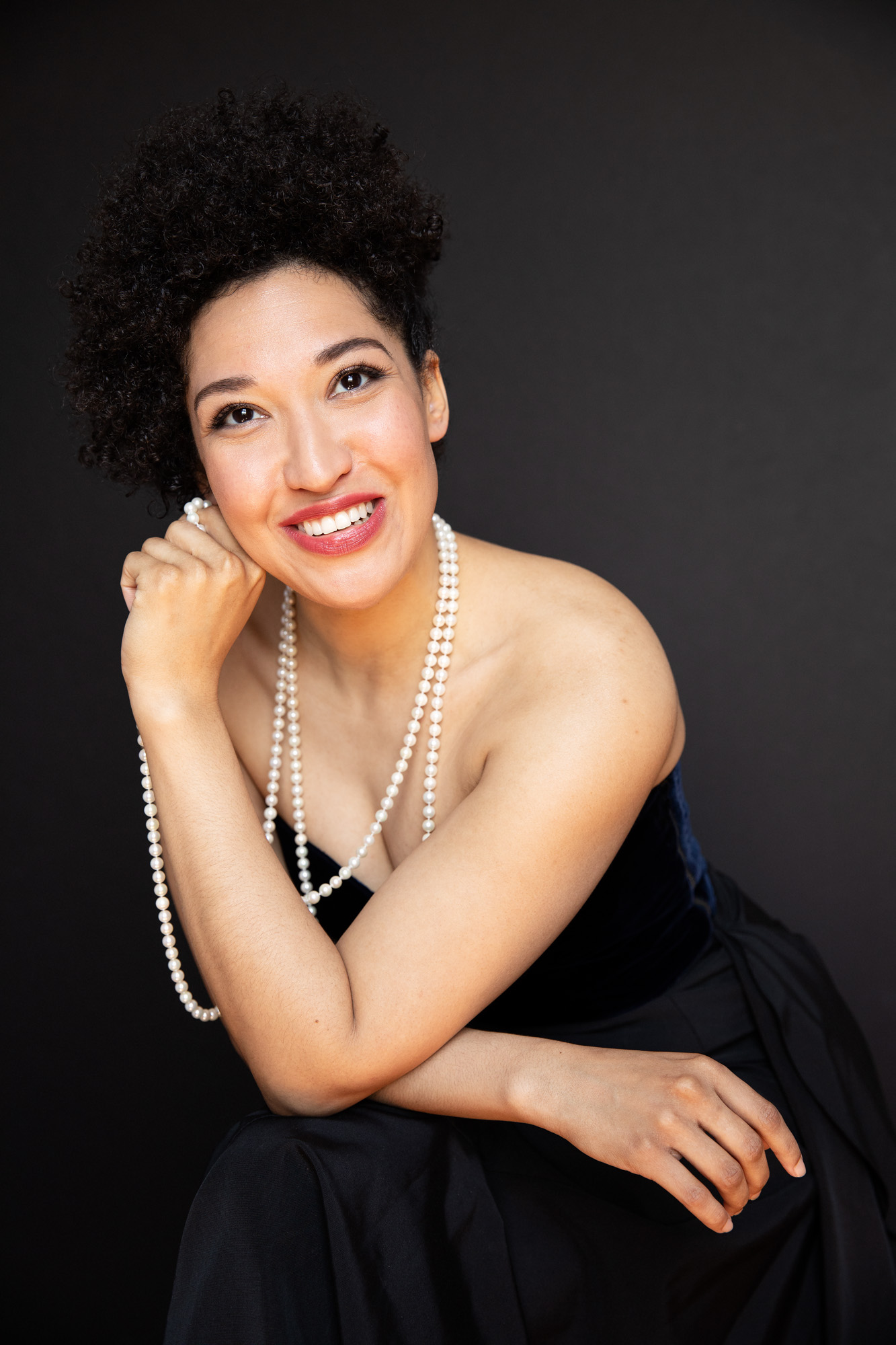 Julia Bullock wears a black sleeveless gown and a white pearl necklace as she leans forward, resting her cheek on her hand and smiling.