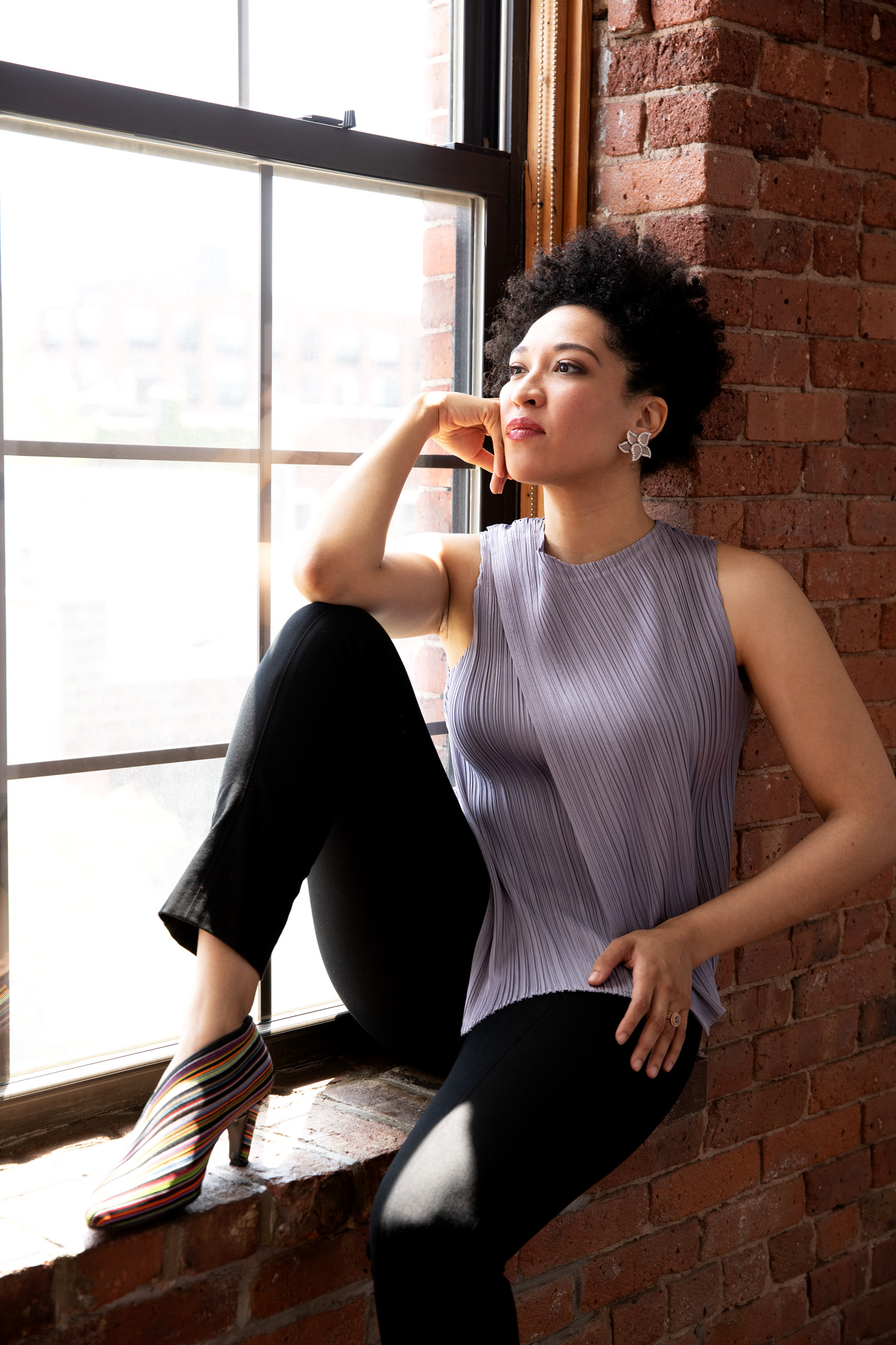 Julia Bullock sits on a red brick window ledge with one elbow resting on her knee, looking pensively and wearing a lavender sleeveless blouse, black tapered pants, and crystal and diamond earrings.