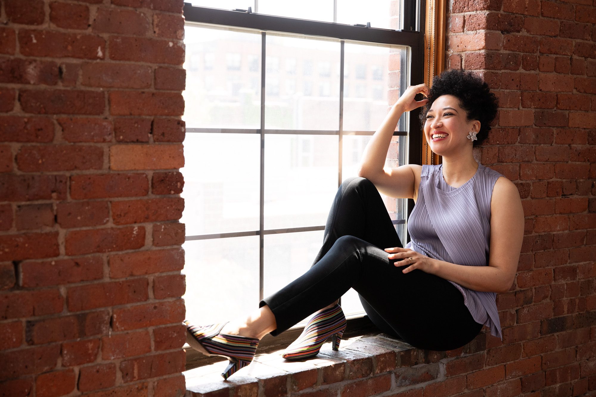 Julia Bullock sits on a red brick window ledge smiling, touching her hair, and wearing a lavender sleeveless blouse, black tapered pants, and crystal and diamond earrings.