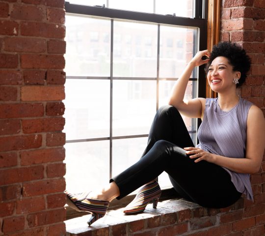 Julia Bullock sits on a red brick window ledge smiling, touching her hair, and wearing a lavender sleeveless blouse, black tapered pants, and crystal and diamond earrings.