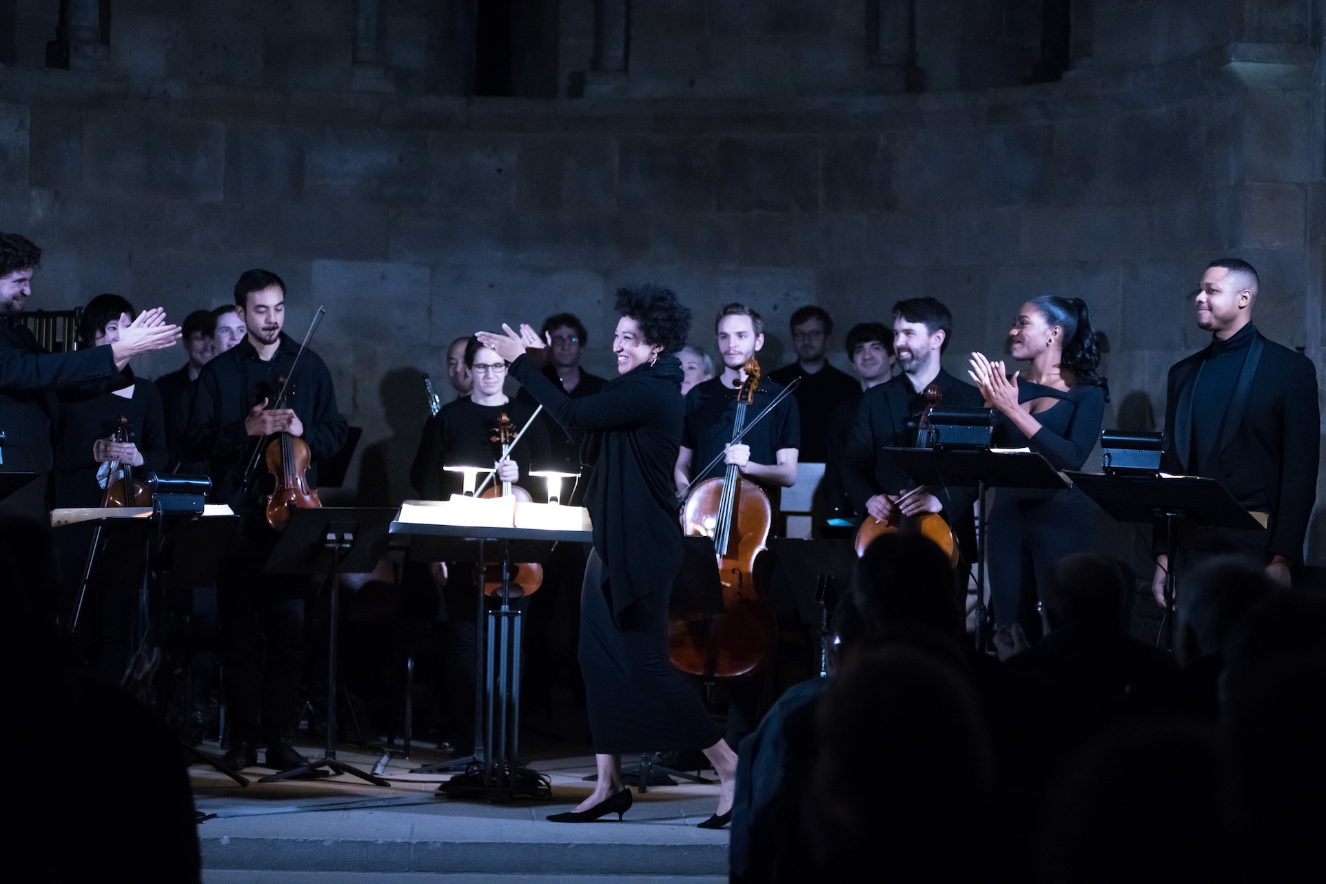 apping for Julia Bullock stands in front of an orchestra in a dark room clthem as they clap for her.