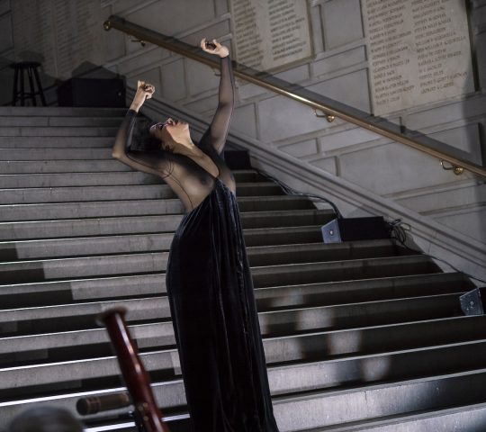 Julia Bullock stands on a stair case wearing a dark gown with a sheer top with her arms extended above her head, which is thrown back.