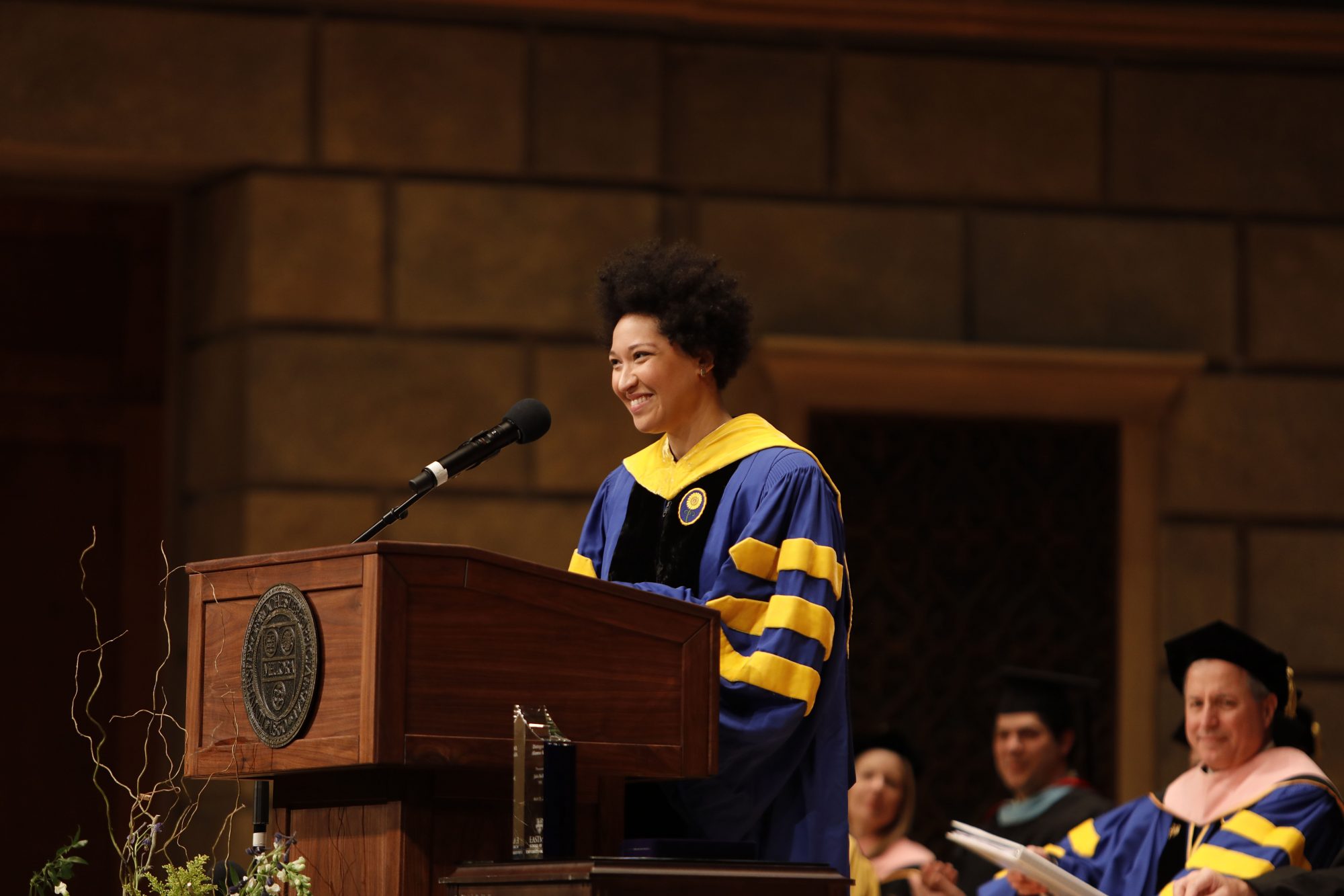 Julia Bullock gives the commencement speech at the Eastman School of Music. She stands at a podium smiling and wearing a graduation gown.