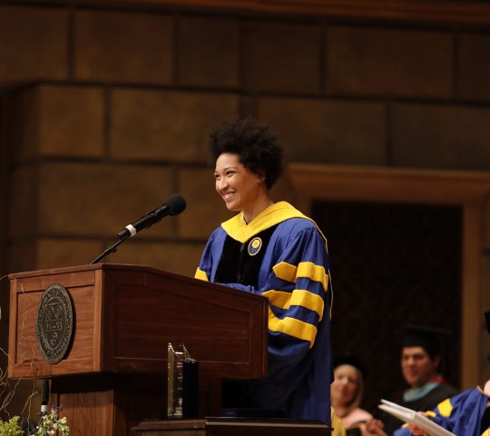 Julia Bullock gives the commencement speech at the Eastman School of Music. She stands at a podium smiling and wearing a graduation gown.