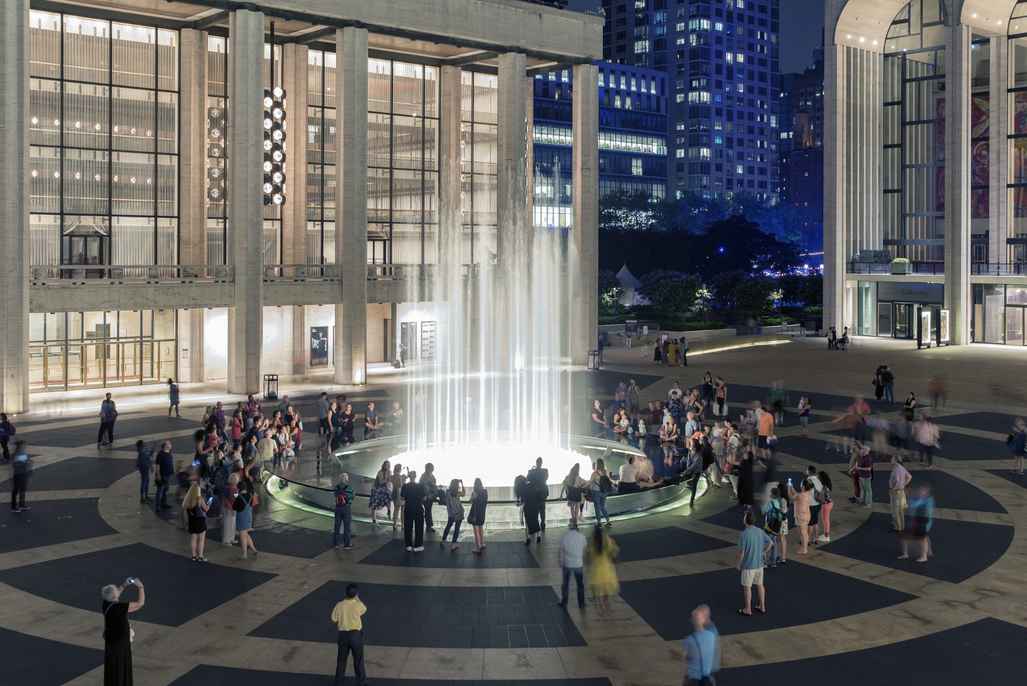 A view of the fountain at Lincoln Center at night with people surrounding the fountain, watching streams of water shoot up.