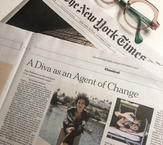 Julia Bullock's New York Times feature titled, "A Diva as an Agent of Change" on a table along with a pair of pink and green glasses.