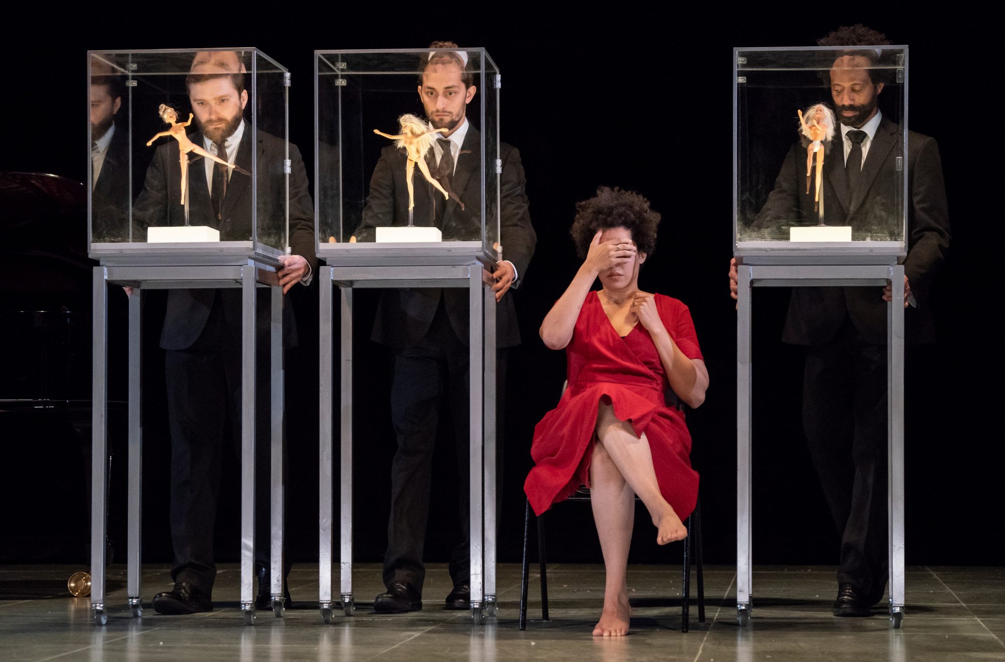 Julia Bullock performing in "Zauberland" at Lincoln Center's White Light Festival. She is wearing a red dress and sitting on a chair, surrounded by three men in suits pushing rolling carts with glass cases that contain naked barbie dolls.