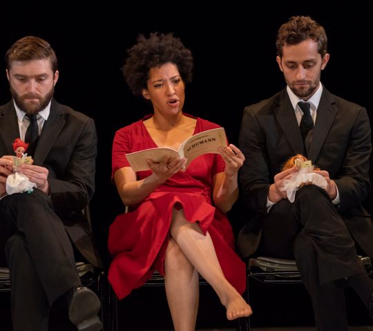 Julia Bullock performing in "Zauberland" at Lincoln Center's White Light Festival. She is wearing a red dress, sitting on a chair, and singing from a score of Schuamann's Dichterliebe, with two men in suits sitting on either side of her holding small dolls.